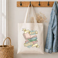 Tote bag with book-themed design hanging on a hook with a denim jacket and basket in the background.